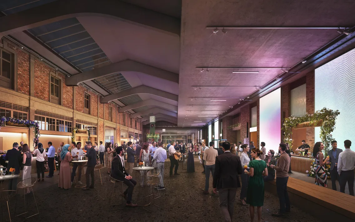 People gather and socialize at a large indoor event space with high ceilings, brick walls, and decorated tables. A bar and stage are visible, with ambient lighting throughout the venue.