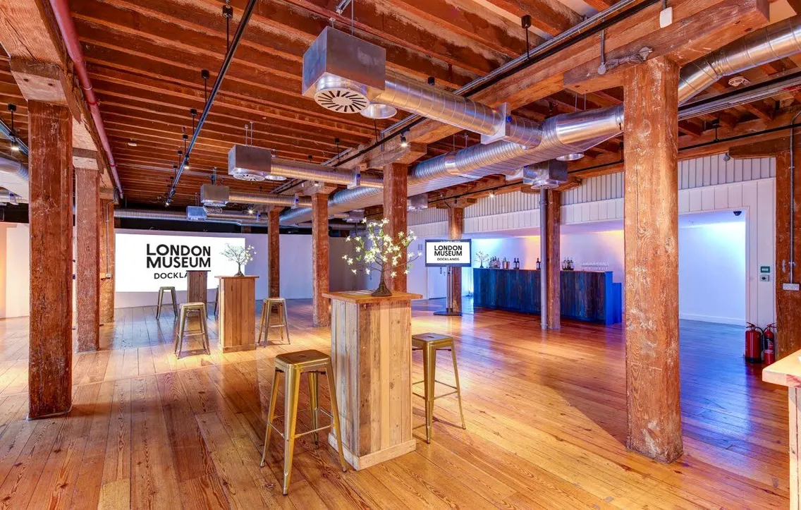 A nearly-empty room with wooden floor, pitch pine columns and exposed brick walls with a screen reading 'London Museum'