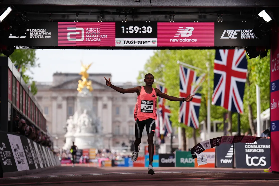 Two people about the cross a red banner as part of a marathon race, with one person waving at a crowd