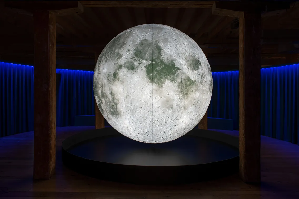 A large illuminated model of the Moon is displayed on a circular platform in a dimly lit room with blue accent lighting.