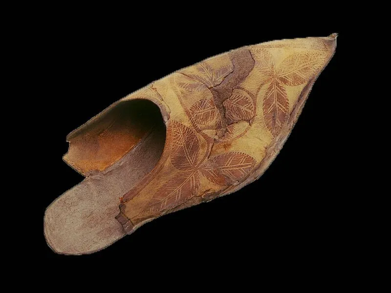 A single, ancient-looking shoe made of organic material with a pointed toe and faded leaf-patterned design.