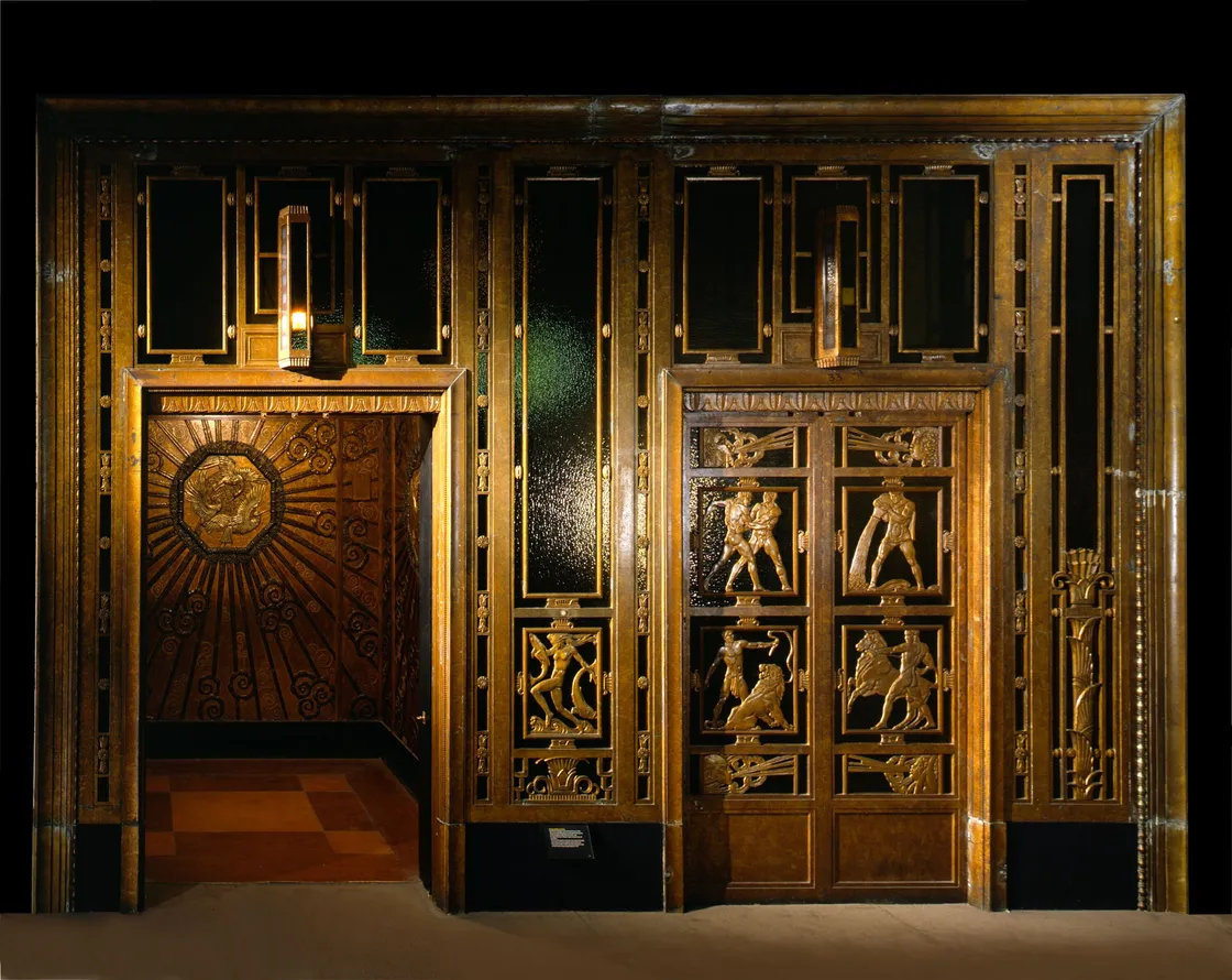 Ornate wooden panel with intricate carvings and a sunburst design on the door, exhibited in a dimly lit setting.