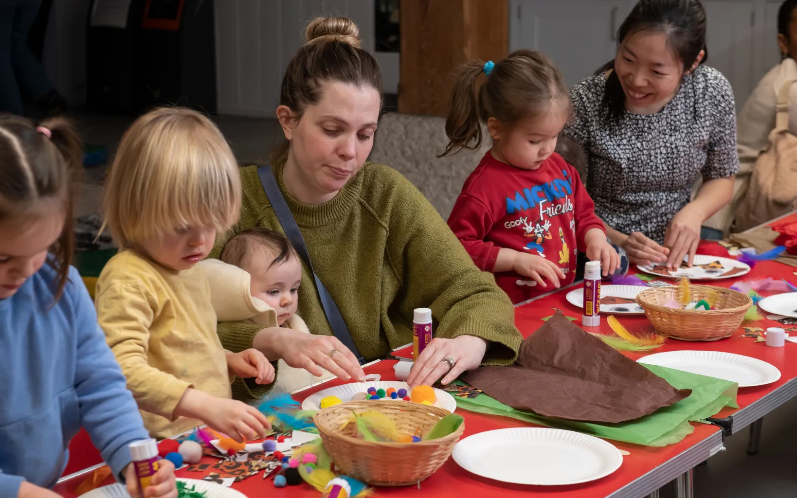 Adults and young children sit at a table engaged in arts and crafts activities with paper, glue, and colorful materials.