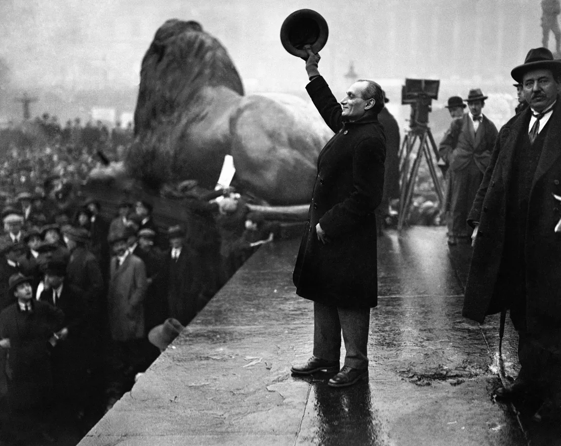 A man stood holding his hat to the sky on a stage in front of a crowd.