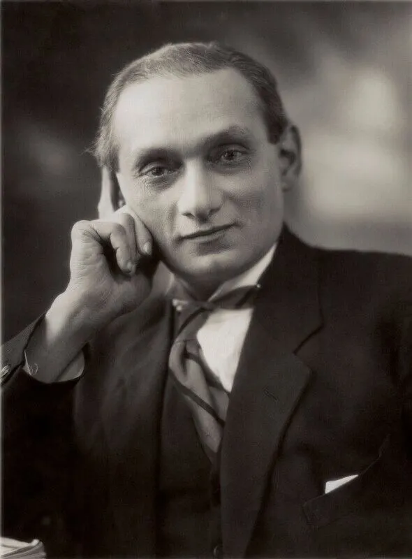 A South Asian man wearing a suit looking directly at the camera, with his hand on his cheek.