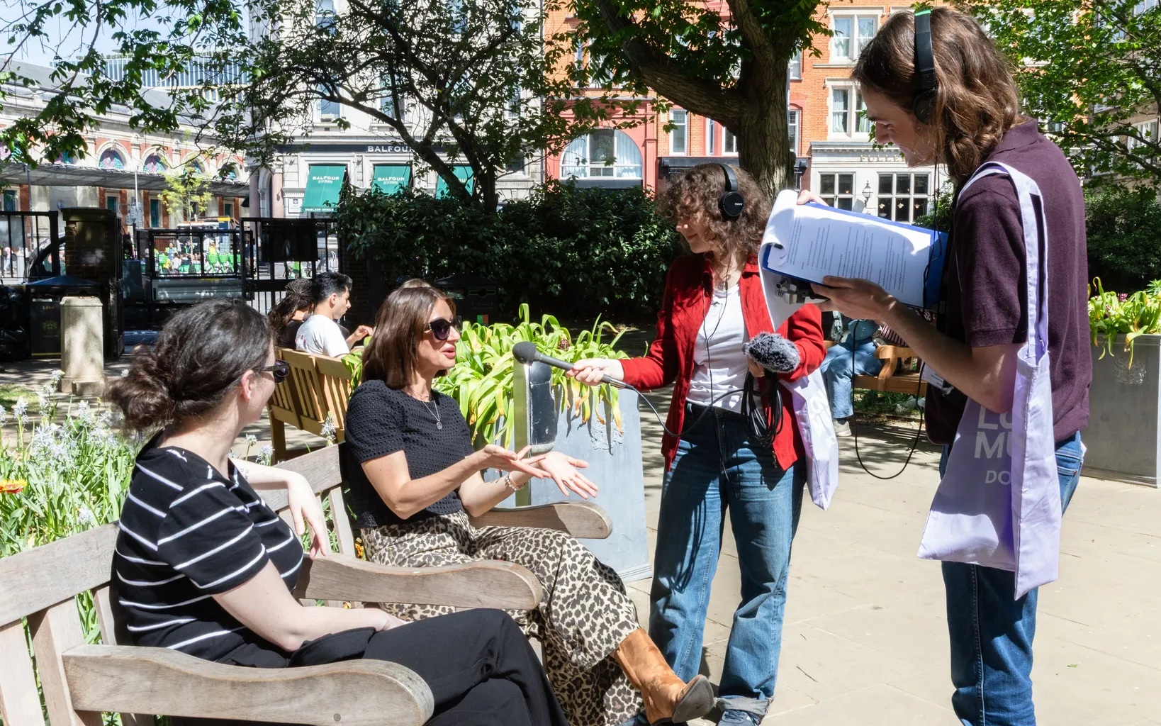 Four people interact outdoors on a sunny day; two are seated on a bench talking, while two others stand nearby, one holding papers and wearing headphones.