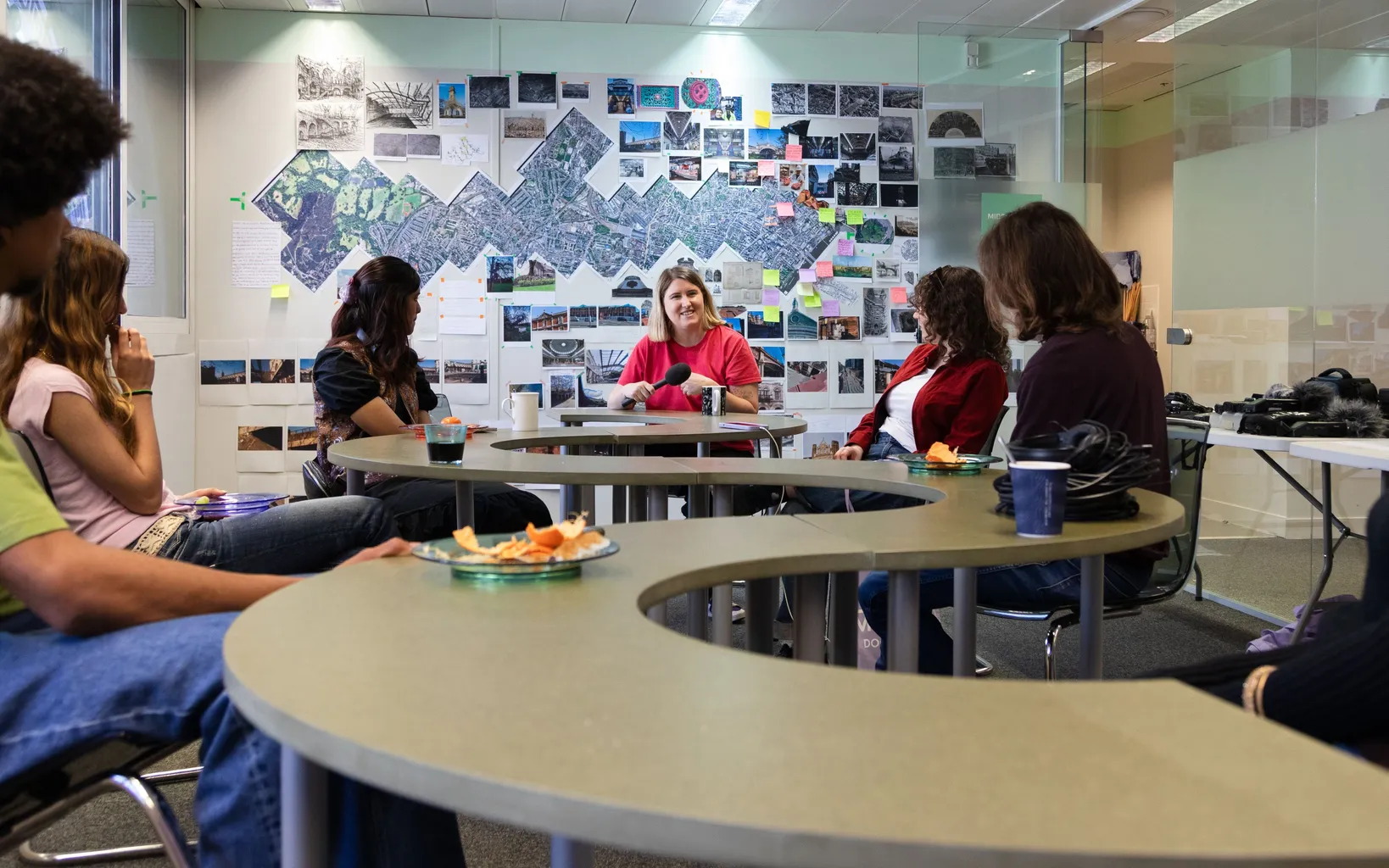 A small group of people sit around curved tables in a meeting room, listening to a woman speaking, with maps and photos displayed on the wall behind her.