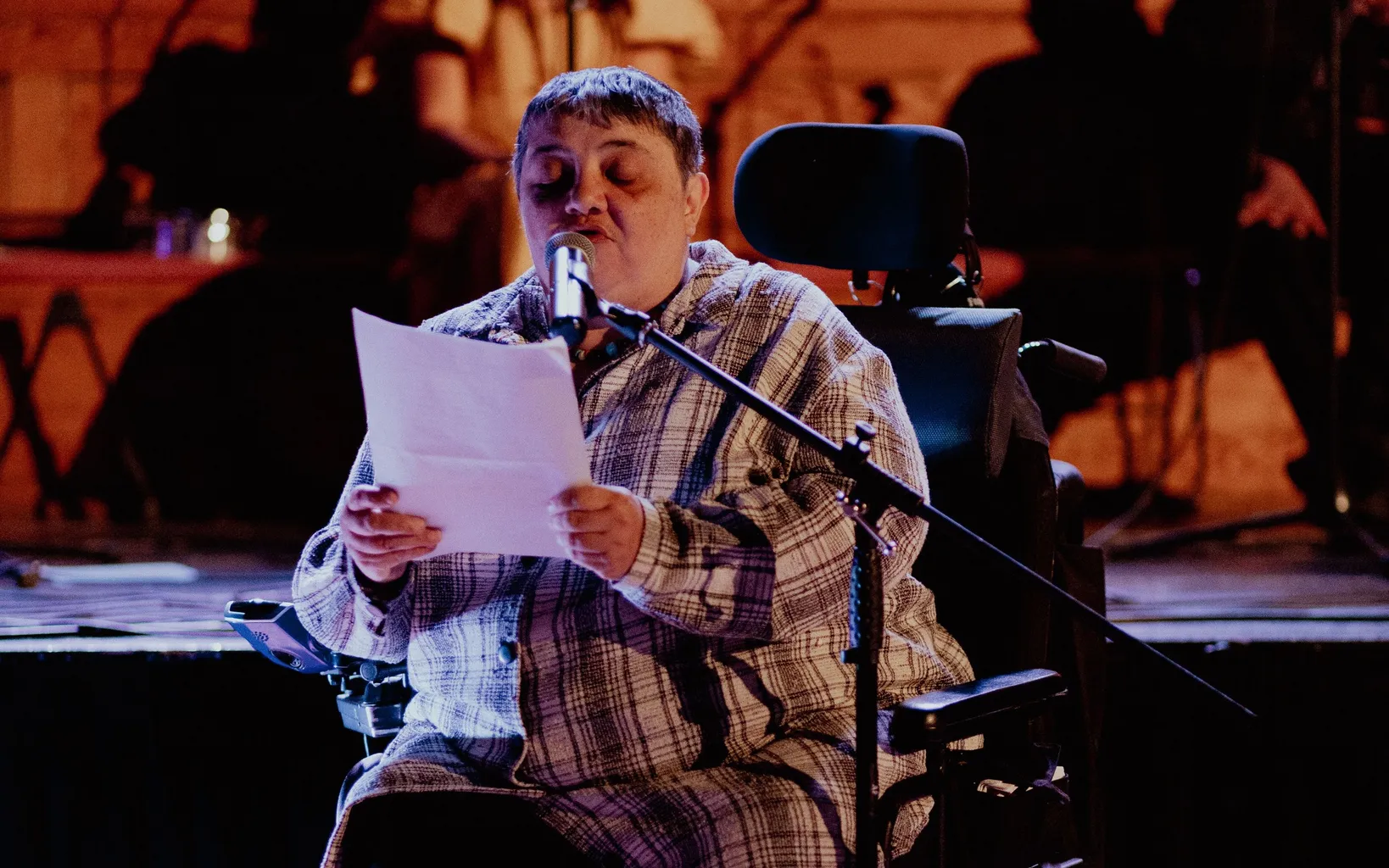 A woman in a wheelchair reads from a piece of paper in front of a microphone on a stage with dim lighting.