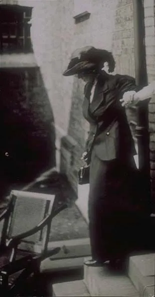 A vintage black-and-white photo of a woman in early 20th-century clothing standing on steps by a building entrance.