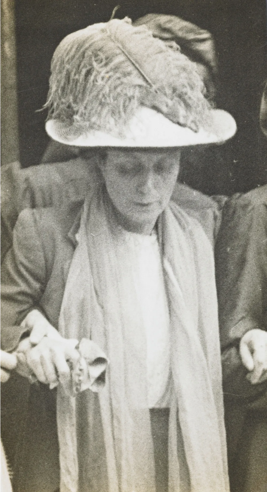 A vintage black-and-white photo of a woman wearing a wide-brimmed hat adorned with feathers, looking down at her hands holding a small object.