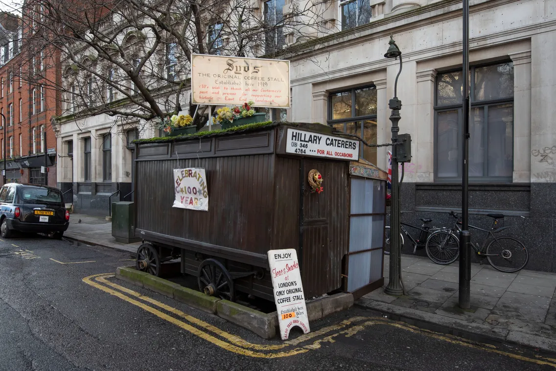 A small wooden food stall on wheels on a street corner, with signage and menu items displayed outside. Bicycles are parked nearby, and a building facade is visible in the background.
