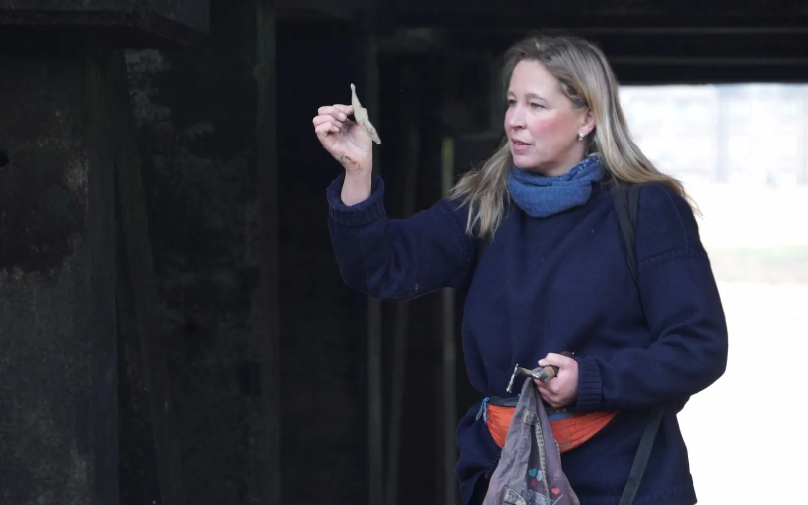 Rusted chain on a pebble-covered riverside with wooden posts and city buildings, including a tall structure, in the background.