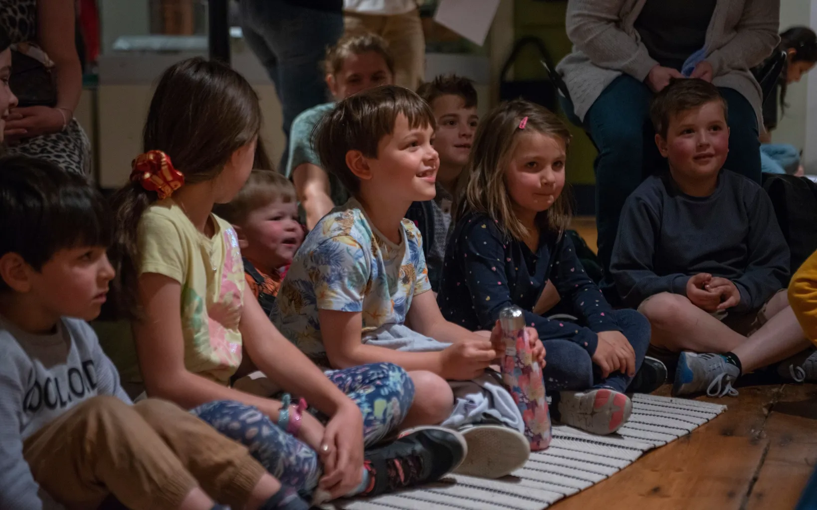 A group of children sit on a rug on the floor, some smiling and engaged, while an adult is partially visible in the background.