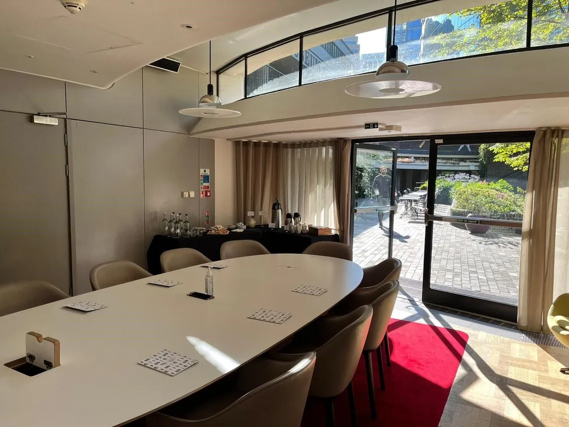 A conference room with an oval table, chairs, and papers. A glass door leads outside. A table with beverages and snacks is near the window. Sunlight illuminates the space.