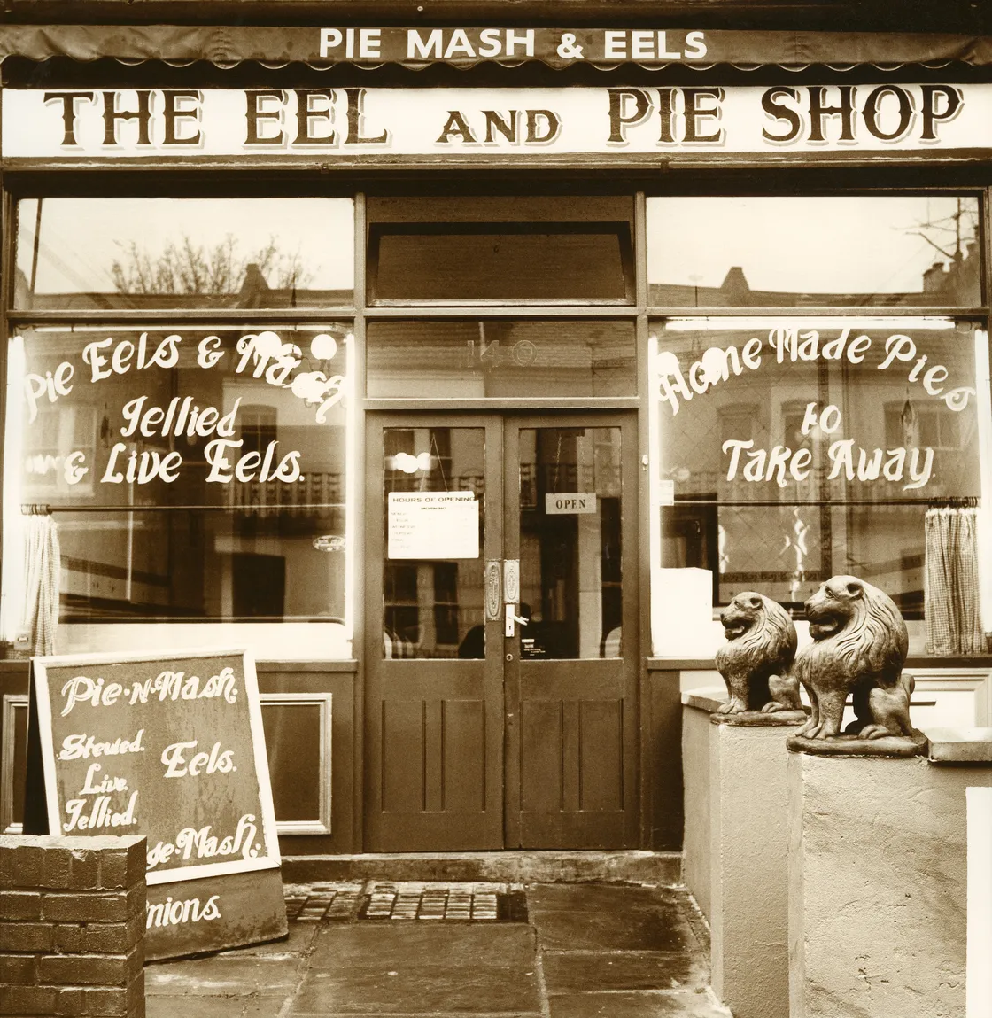 Vintage photo of a traditional british pie and mash shop, with signage for eels and pies, featuring stylized statues of eagles at the entrance.