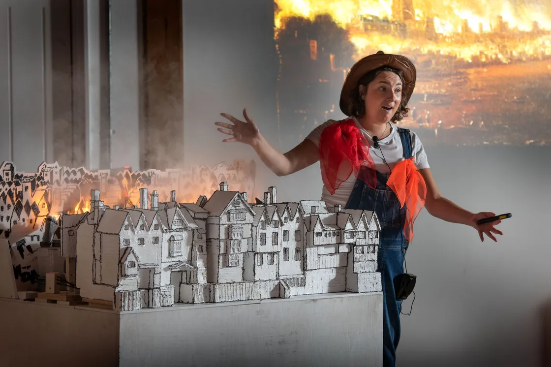 A woman in costume gestures while presenting a model of historic buildings, with smoke and a burning city scene projected in the background.