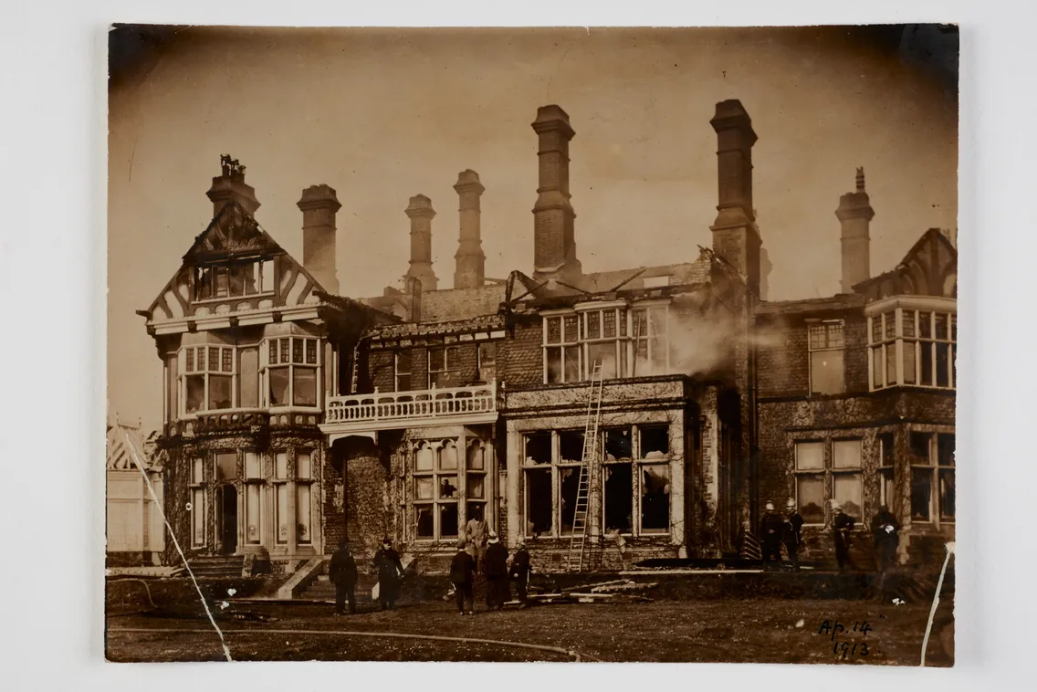 Old black and white photo of a damaged large house with multiple chimneys and people standing in front, dated april 1912.