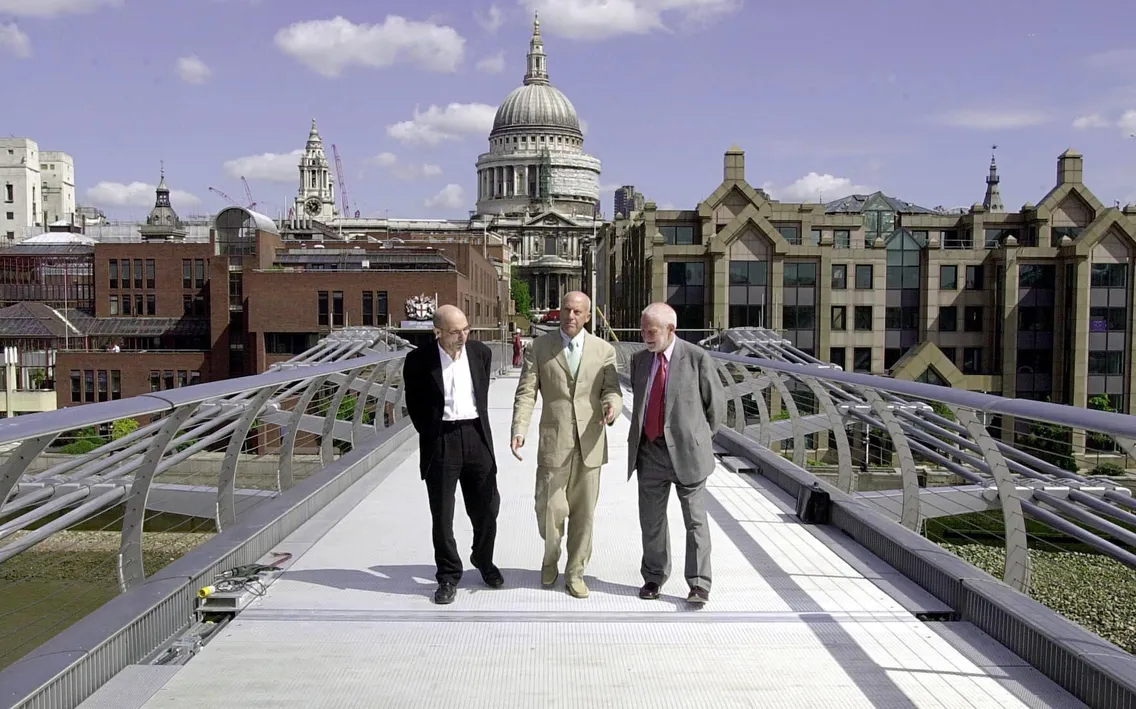Three men in suits walk along the Millennium Bridge in London, with St. Paul’s Cathedral and city buildings in the background under a partly cloudy sky.