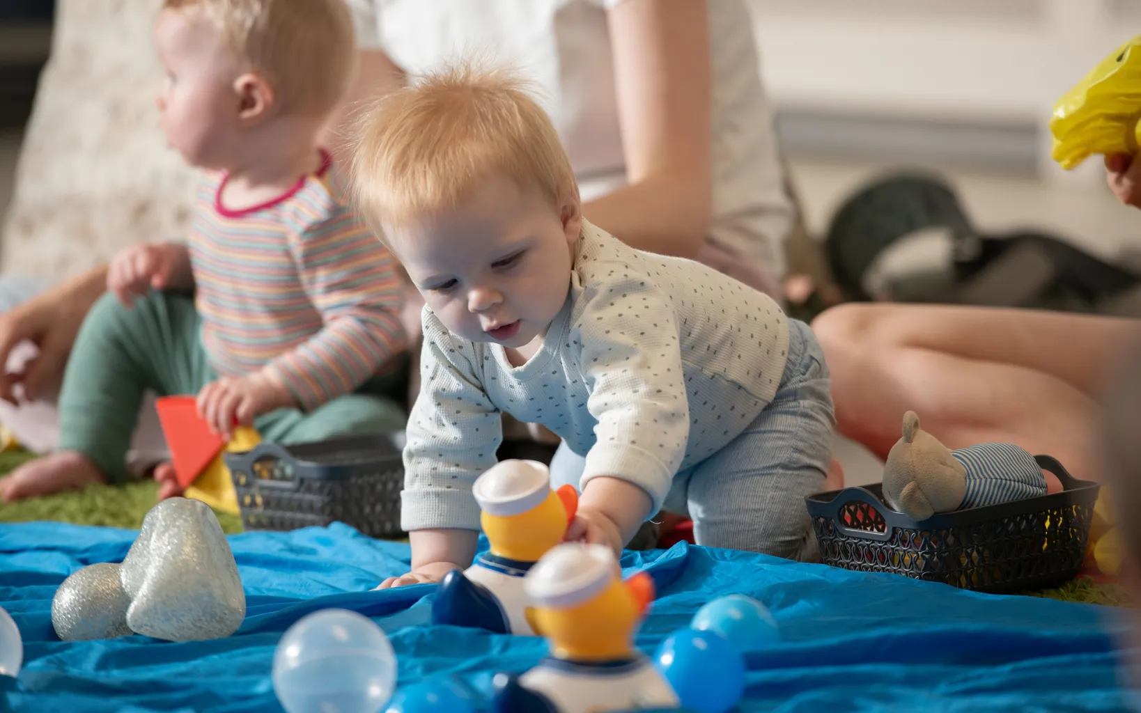 Two babies play on a blue blanket with plastic ducks, while adults sit nearby in the background.