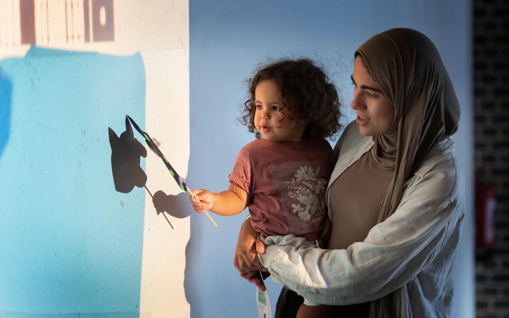 An adult holds a young child who uses a paintbrush to create a shadow of a cat on a wall, with sunlight casting shadows in the background.