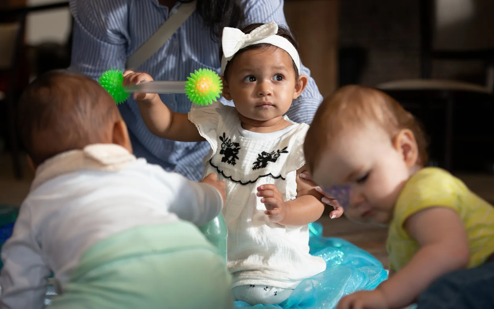 Three toddlers play on the floor, with one holding a green and yellow spiky stick toy, while an adult sits behind them.