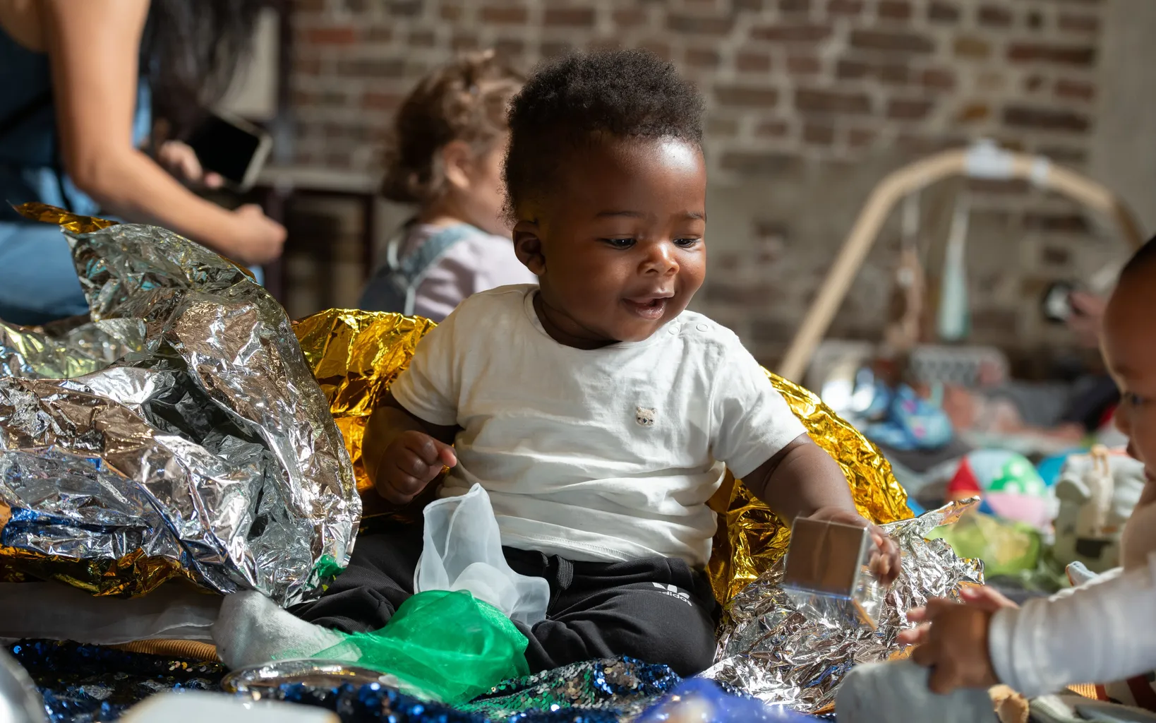 A baby sits on the floor surrounded by shiny foil and toys, interacting with another child nearby.
