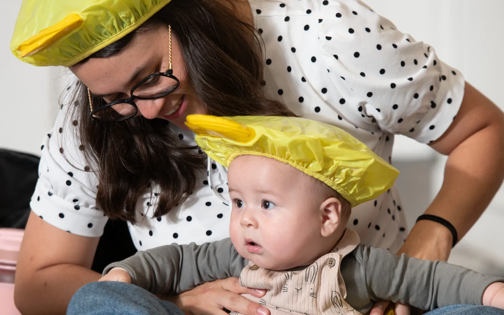 An adult and a baby, both wearing yellow hats and sitting on the floor, play together among blue and yellow toys.