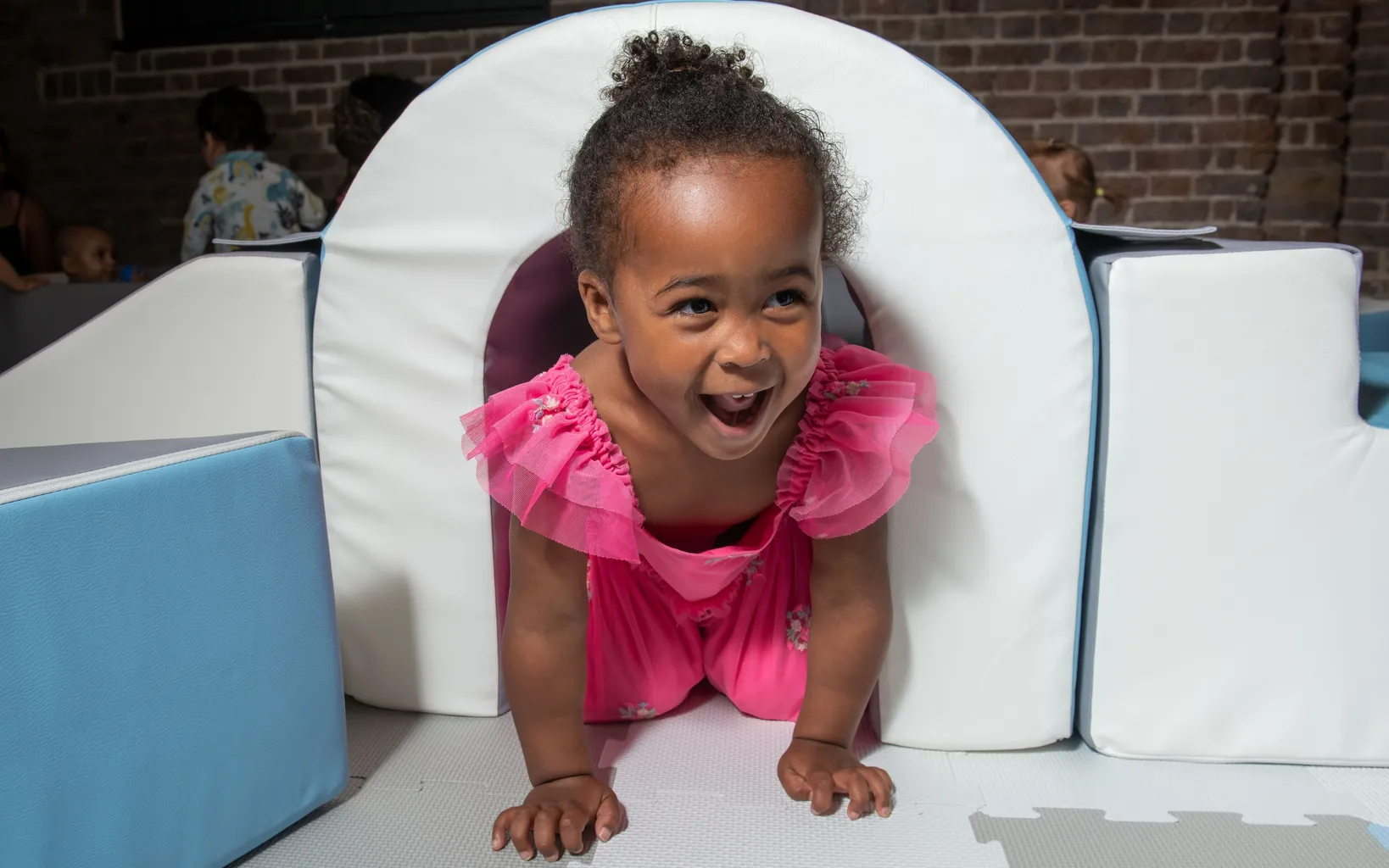 A toddler girl in a pink dress crawls through a white soft play arch.