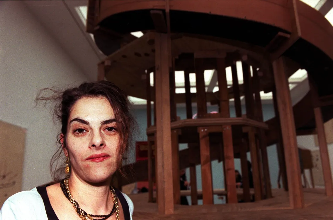 A woman with dark hair stands in front of a large wooden sculpture or architectural model at an exhibition in an indoor setting, the White walls behind her providing a striking backdrop.
