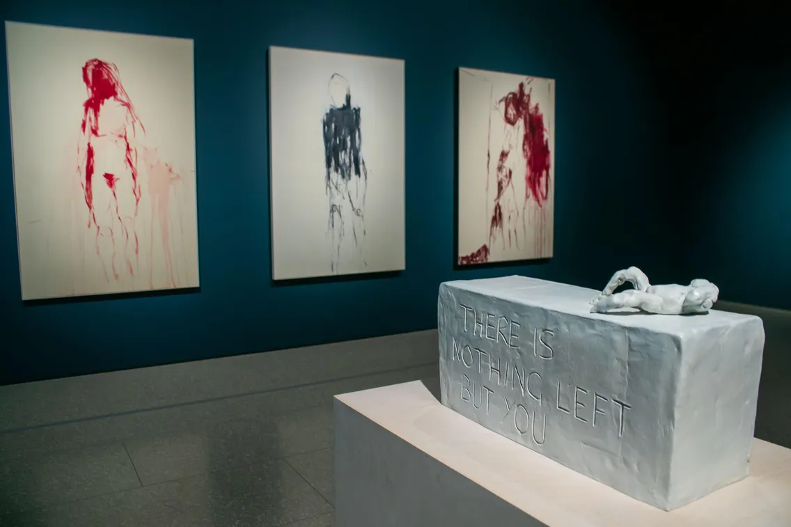 A sculpture of a reclining figure on a block inscribed with "THERE IS NOTHING LEFT BUT YOU," evocative of loneliness, stands before three abstract figure paintings on a blue wall—creating an exhibition atmosphere reminiscent of Tracey's contemplative