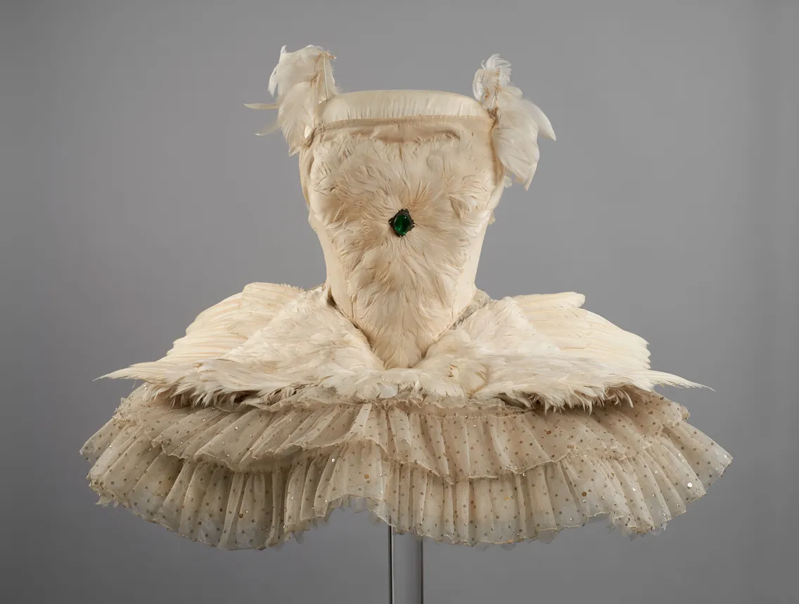 An ornate ballet costume featuring a feathered bodice with a central green gem and a multi-layered, speckled tulle skirt.