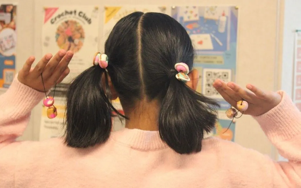 A person with short pigtails decorated with colorful hair ties holds up both hands with beaded strings, facing away from the camera. Posters and an untitled design are visible on the wall in the background.