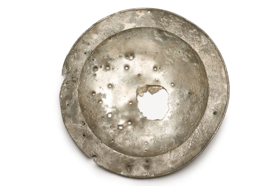 A weathered, circular metal object with a large hole in the center and multiple smaller holes scattered across the surface.