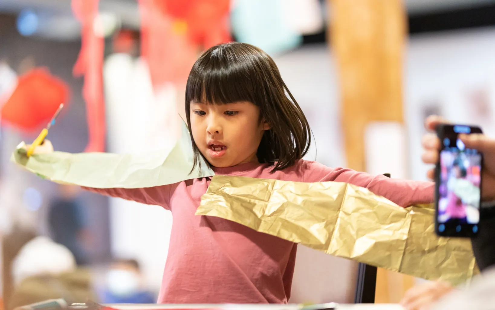 A young girl in a pink shirt plays with colorful paper crafts, extending her arms out while a smartphone captures the moment.
