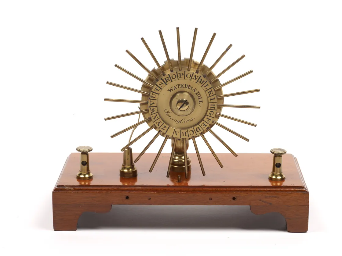 Antique brass and wood telegraph machine with a circular alphabetic dial and radial spokes on a stand, isolated on a white background.