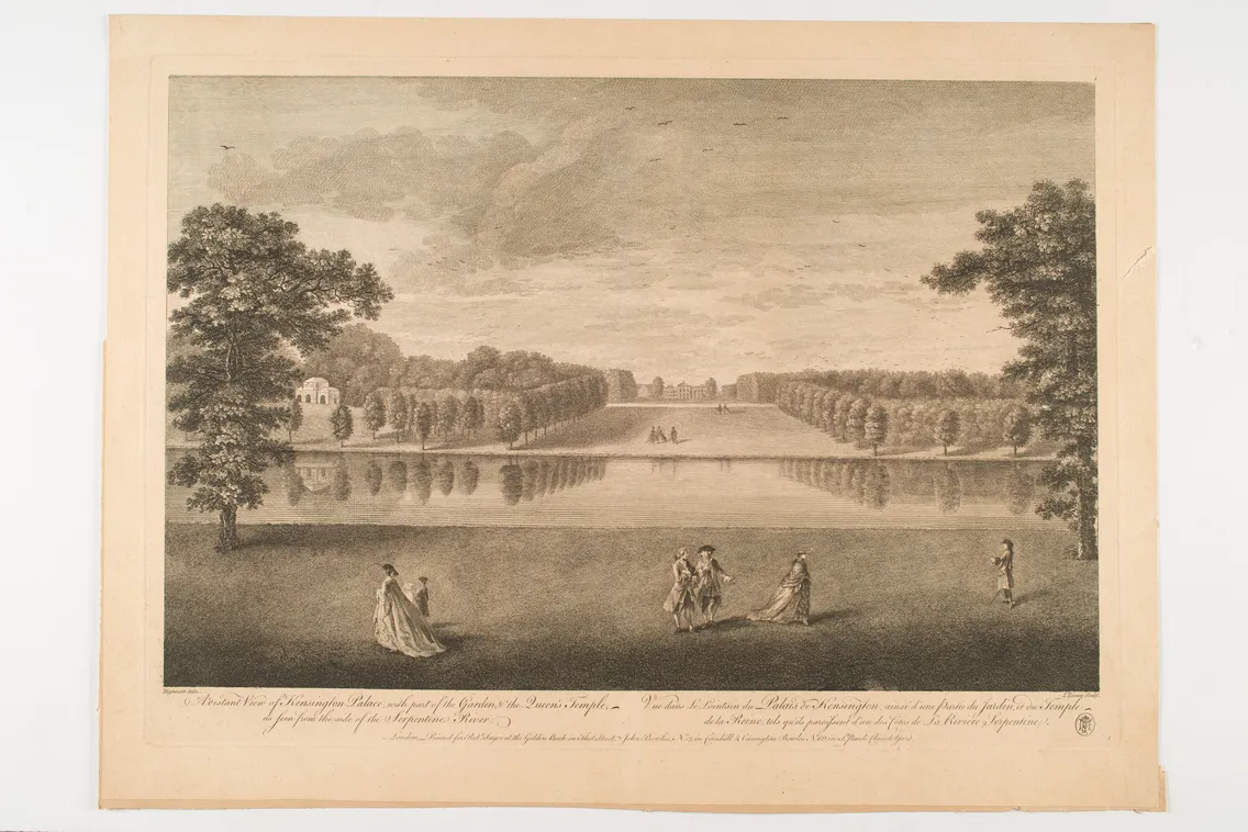 A few people standing on grass in front of a large lake with bushes in the background