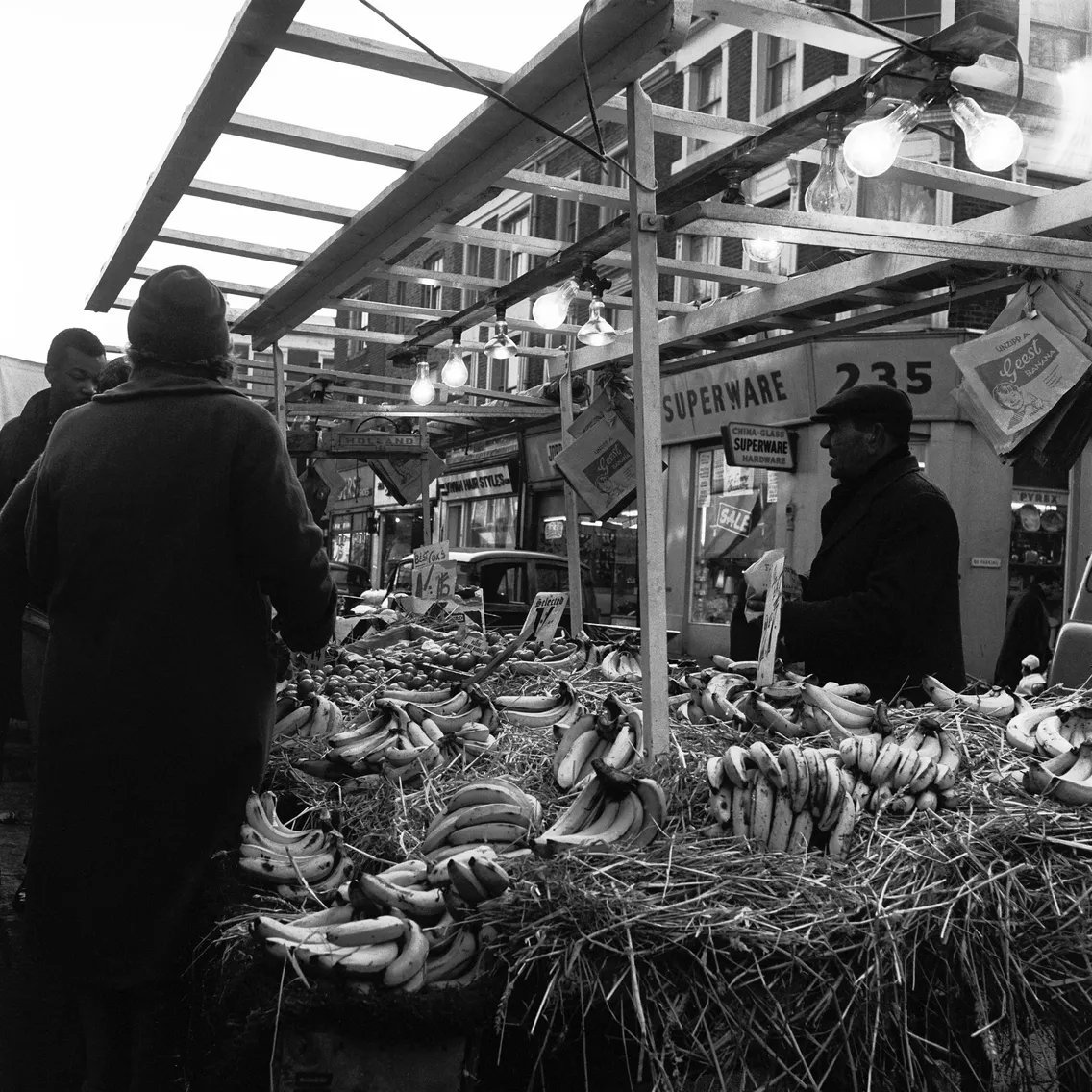 Black and white photo of a bustling outdoor market with stalls displaying bananas under a sign labeled "superware," with customers and vendors interacting.