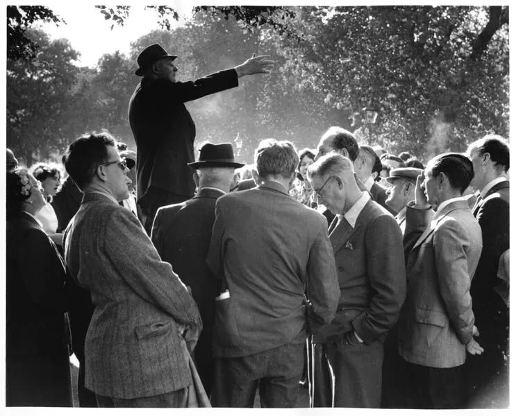 Black and white photo of a man in a hat speaking animatedly to a gathered crowd in a park, with sunlight filtering through trees.
