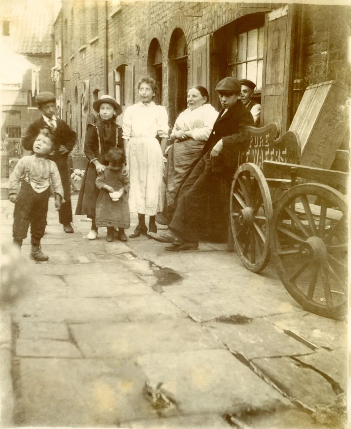 People of varying ages gathering on a cobbled street with old buildings, a child pulling another in a small cart.