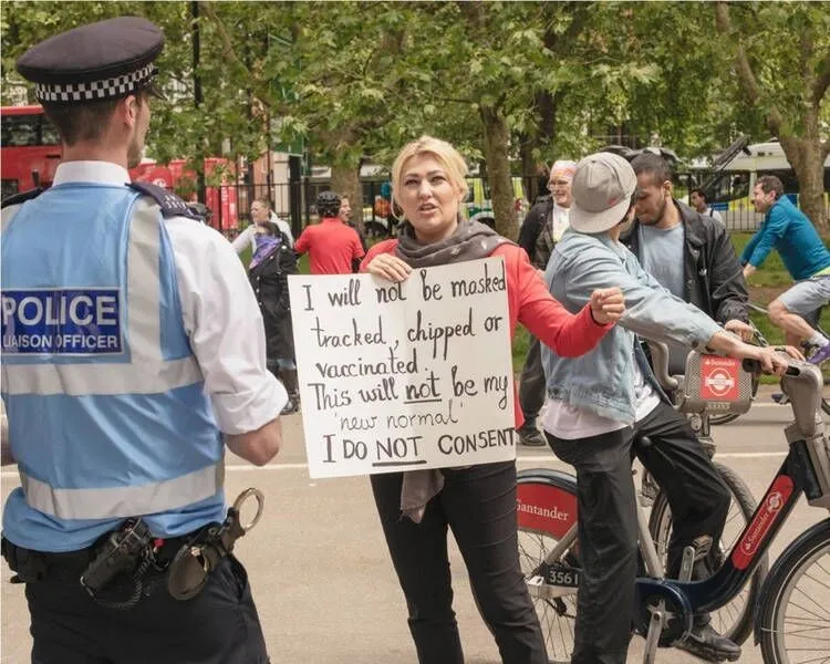 A woman holds a protest sign against tracking and vaccination as she talks to a police officer in a park, with other people around including a cyclist.