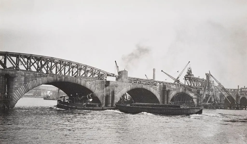 Historical black and white photo of a bridge under construction with arches, a crane, and a boat passing underneath.