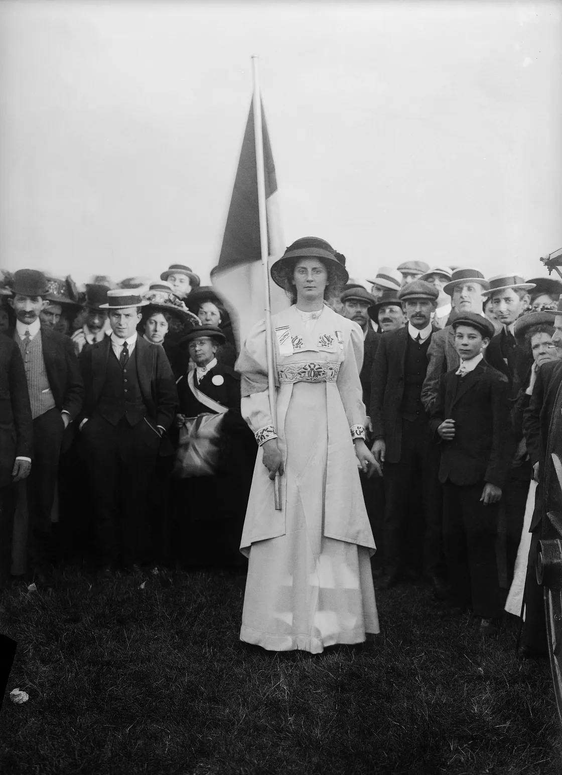 A black and white historical photo of a woman holding a flag, standing in front of a large group of people, mostly men, in an outdoor setting.