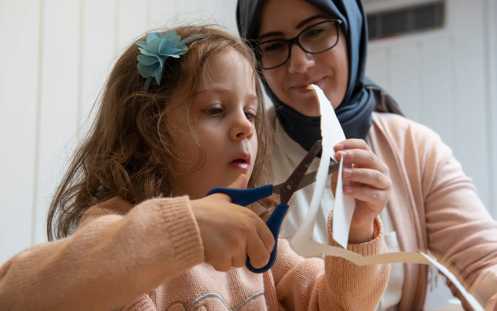 A young girl and a woman in a hijab work together cutting paper with scissors at a table, focused on their craft project.
