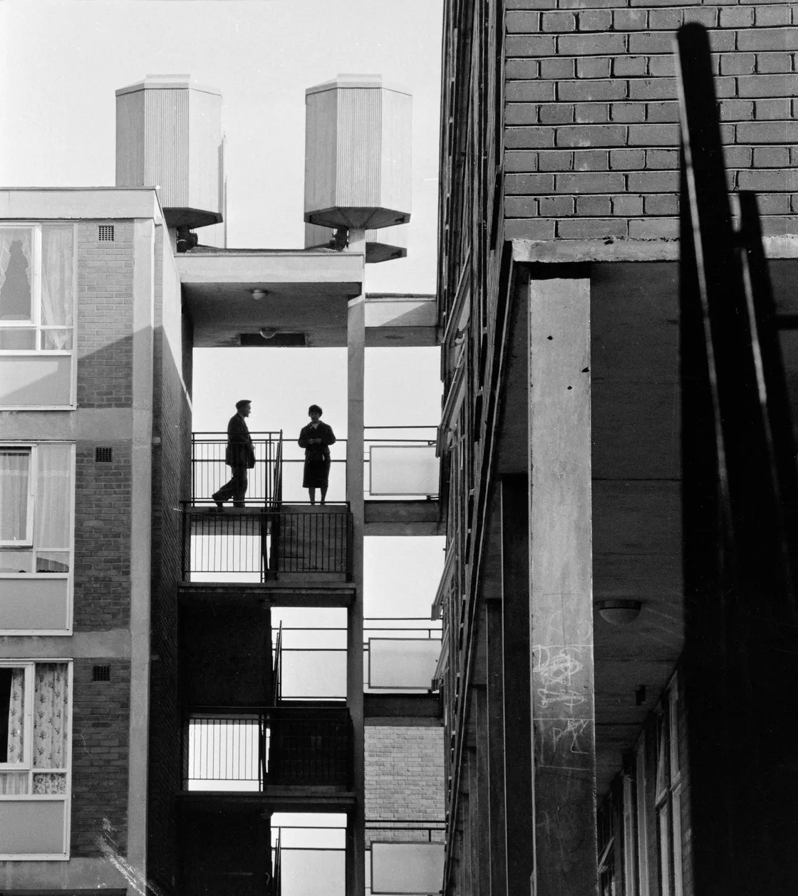 Two people stand on separate balconies of a stark, urban building with large rooftop vents visible above.