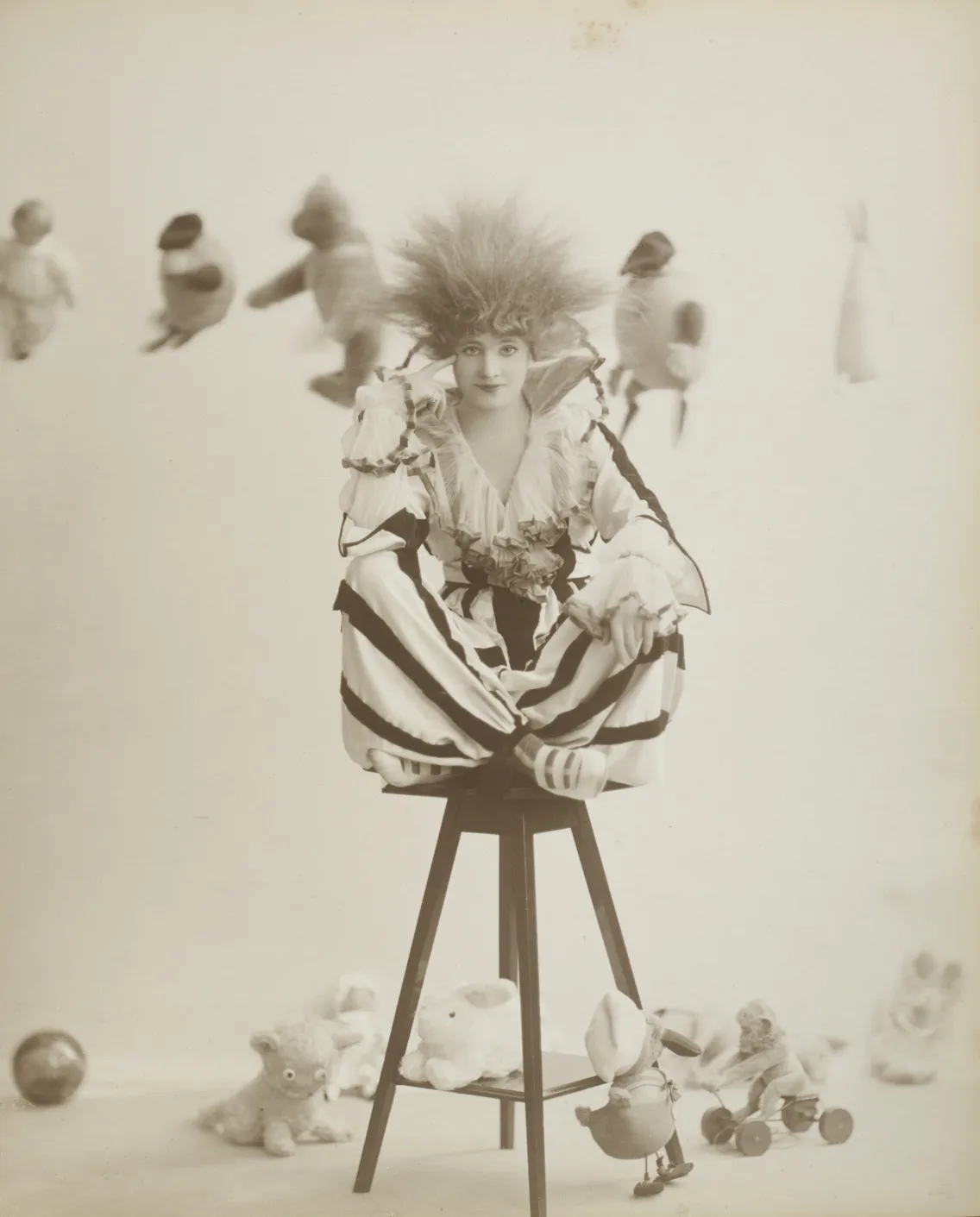 A vintage photograph of a woman seated on a tall stool, surrounded by toys and stuffed animals, wearing a striped costume and a large feathered hat.
