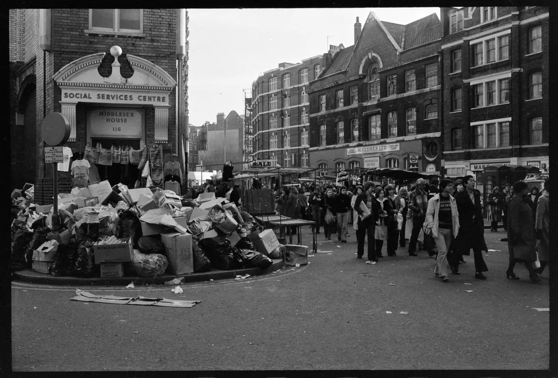 Black and white street scene with a large pile of garbage in front of a social services centre, with pedestrians walking by in a busy urban area.