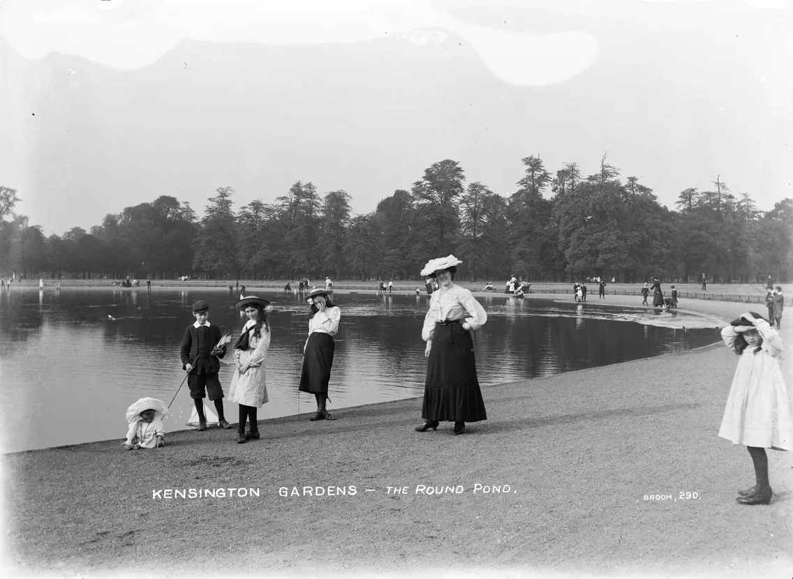 Vintage black-and-white photo of people in early 20th-century clothing standing by the round pond in kensington gardens.