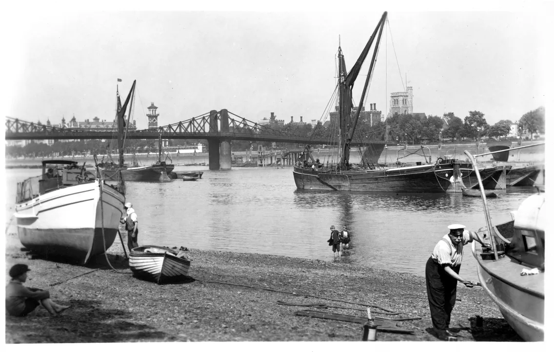 Historic black and white photograph showing boats on the river thames with a bridge and buildings in the background. some individuals are visible near the water's edge.