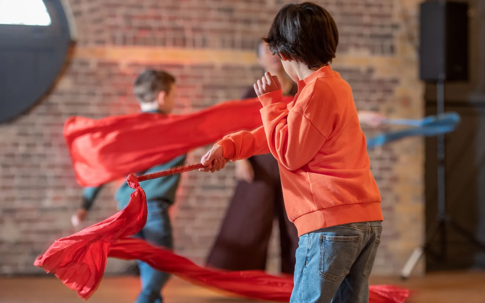 Four children stand dotted around a wooden floor with their backs turned to the camera. In the foreground stands a boy with a blue and red striped top. His right arm swings out a long pink fabric ribbon. The others are out of focus, but do the same.