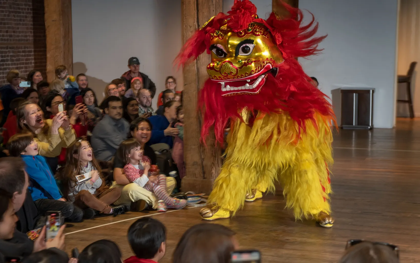 A crowd of adults and children sit on a wooden floor as they watch a traditional red and gold lion puppet spring forward.